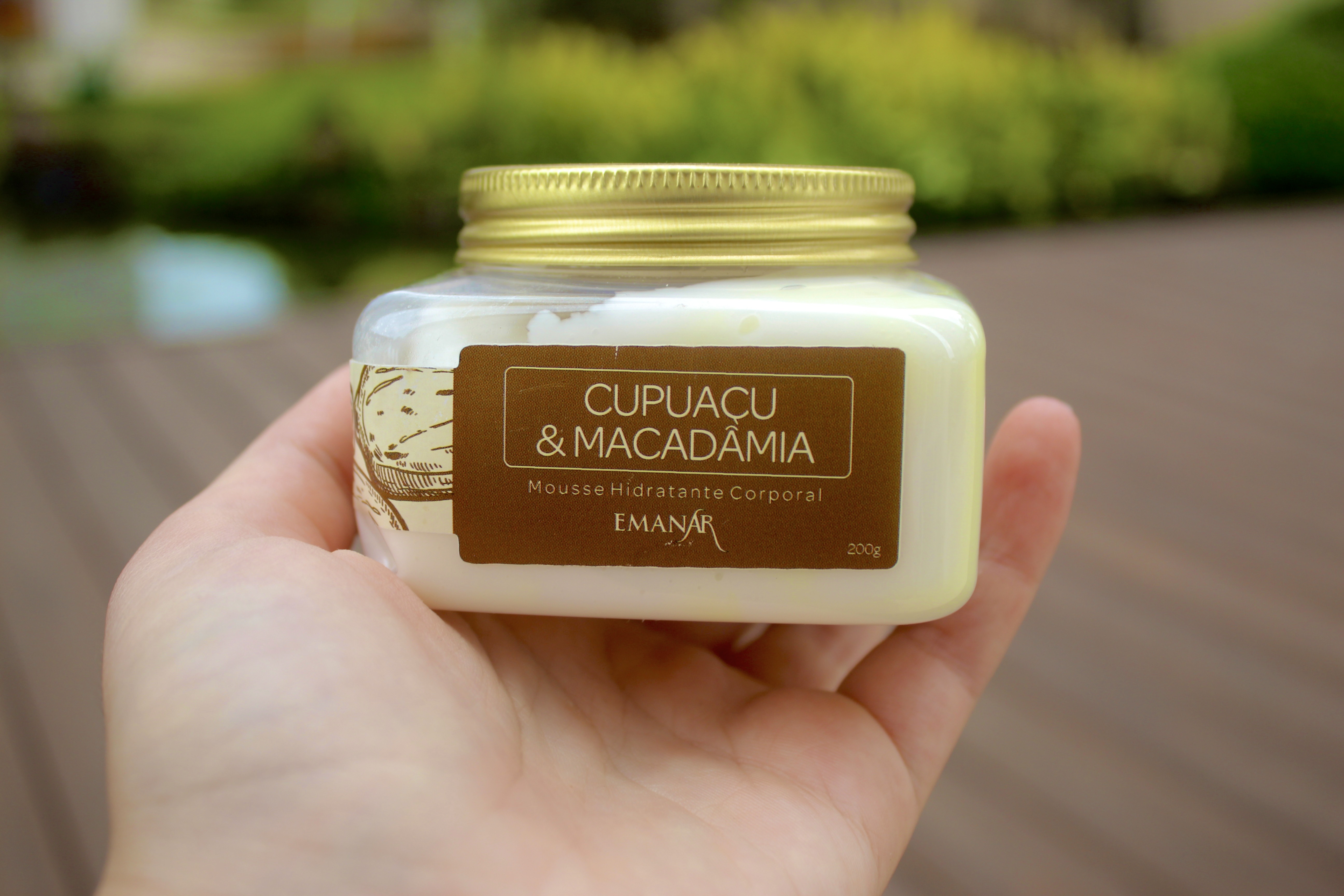 Frasco de mousse hidratante corporal com rótulo 'CUPUAÇU & MACADÂMIA', segurado por uma mão, fundo desfocado de um ambiente natural.